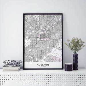 Puede incluir: Un mapa en blanco y negro de Adelaida, Australia, impreso en papel blanco y enmarcado en un marco negro. El mapa se muestra en un estante blanco con un cuenco de metal negro y una pila de libros.
