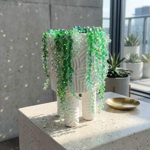 May include: White ceramic planter with a textured design, supported by three legs. The planter is adorned with cascading strands of green and clear beads, resembling a trailing plant. A small gold dish sits nearby.