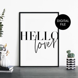 Puede incluir: Arte de pared imprimible en blanco y negro con el texto "HELLO lover" en una fuente moderna. El arte está enmarcado en un marco negro y se muestra en una mesa con un jarrón y una planta en maceta.