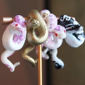 Peut inclure: Quatre boucles d'oreilles en forme de serpent sur un support doré. Les boucles d'oreilles sont en résine blanche, noire et rose et ont un design de serpent réaliste.