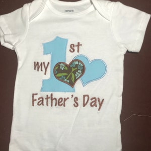 K&ouml;nnte beinhalten: Wei&szlig;er Baby-Strampler mit blauer und brauner Stickerei. Die Stickerei lautet "My 1st Father's Day" mit einer gro&szlig;en blauen Zahl eins und einem Herzen.