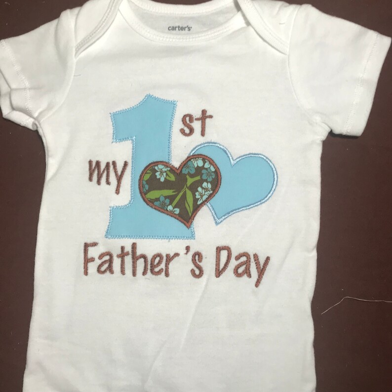K&ouml;nnte beinhalten: Wei&szlig;er Baby-Strampler mit blauer und brauner Stickerei. Die Stickerei lautet "My 1st Father's Day" mit einer gro&szlig;en blauen Zahl eins und einem Herzen.