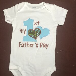K&ouml;nnte beinhalten: Wei&szlig;er Baby-Strampler mit blauer und brauner Stickerei. Die Stickerei lautet "My 1st Father's Day" mit einem blauen Herzen und einem braunen Herzen mit Blumenstickerei.