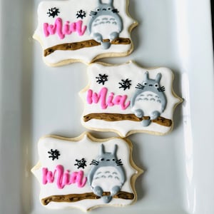 May include: Three decorated sugar cookies with a gray and white cartoon character, a brown branch, and black spiders. The cookies are decorated with pink icing and the name "Mia" is written on each cookie.