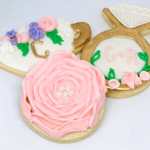 May include: Three decorated sugar cookies. One cookie is shaped like a teacup with pink and purple flowers and the letter "C" in gold. Another cookie is shaped like a ring with a gold band and a white diamond. The third cookie is a pink rose with white pearl accents. The cookies are decorated with green leaves and pink flowers.