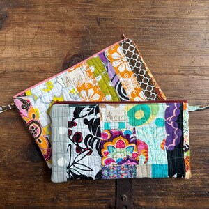 May include: Two quilted fabric pouches with zipper closures. The top pouch has a pink zipper and a floral pattern with orange, yellow, and white flowers. The bottom pouch features a patchwork design with various colors and patterns, including floral and geometric shapes.