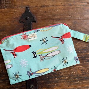 May include: A light blue zippered pouch with a whimsical pattern of red, yellow, and gray stylized creatures and snowflakes. The pouch has a red zipper and a small fabric tag that says "Alala". A matching fabric handle is attached.