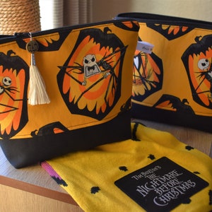 May include: Two yellow and black zippered pouches with a black and white Jack Skellington print. The pouches have a black tassel and a metal charm. A yellow and black fabric with the text "Tim Burton's The Nightmare Before Christmas" is in the foreground.