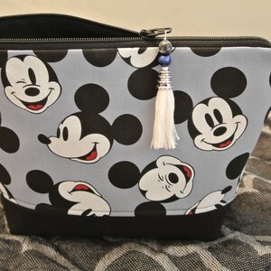 May include: A grey zippered pouch with a black trim and a white tassel. The pouch is decorated with a repeating pattern of Mickey Mouse heads in black and white.