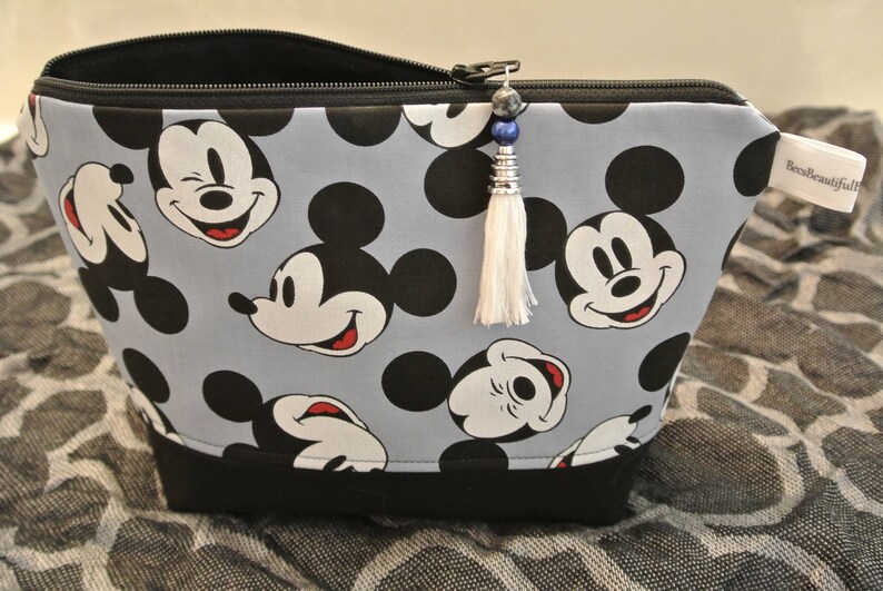 May include: A grey zippered pouch with a black trim and a white tassel. The pouch is decorated with a repeating pattern of Mickey Mouse heads in black and white.