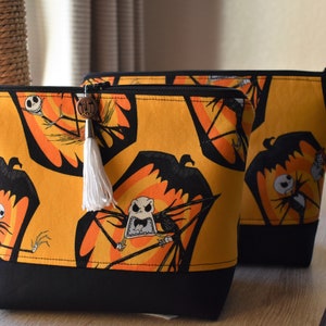 May include: Two zippered pouches with a black and orange fabric featuring a repeated pattern of Jack Skellington from The Nightmare Before Christmas. The larger pouch has a white tassel.