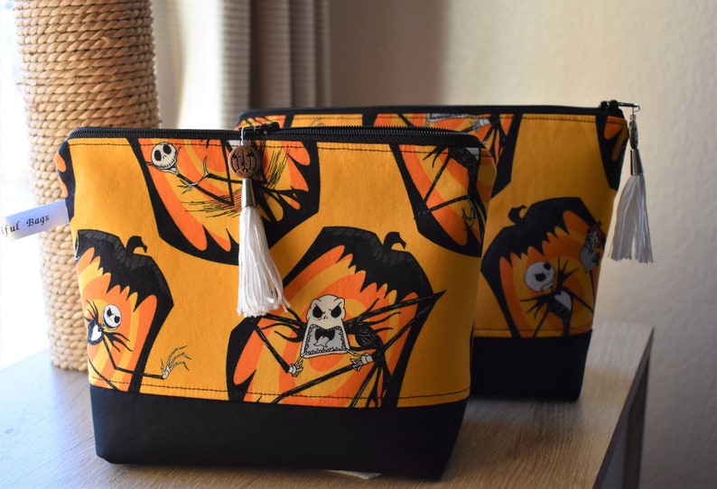 May include: Two zippered pouches with a black and orange fabric featuring a repeated pattern of Jack Skellington from The Nightmare Before Christmas. The larger pouch has a white tassel.