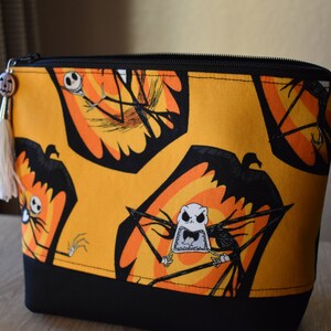 May include: A yellow zippered pouch with a black bottom. The pouch is decorated with a repeating pattern of Jack Skellington from The Nightmare Before Christmas. The pattern features Jack Skellington's head and body in a black silhouette with orange and yellow accents.