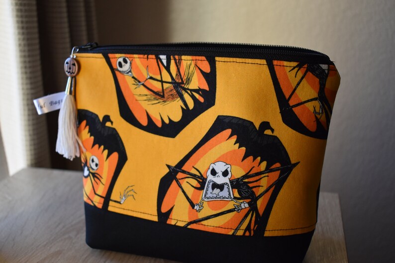 May include: A yellow zippered pouch with a black bottom. The pouch is decorated with a repeating pattern of Jack Skellington from The Nightmare Before Christmas. The pattern features Jack Skellington's head and body in a black silhouette with orange and yellow accents.