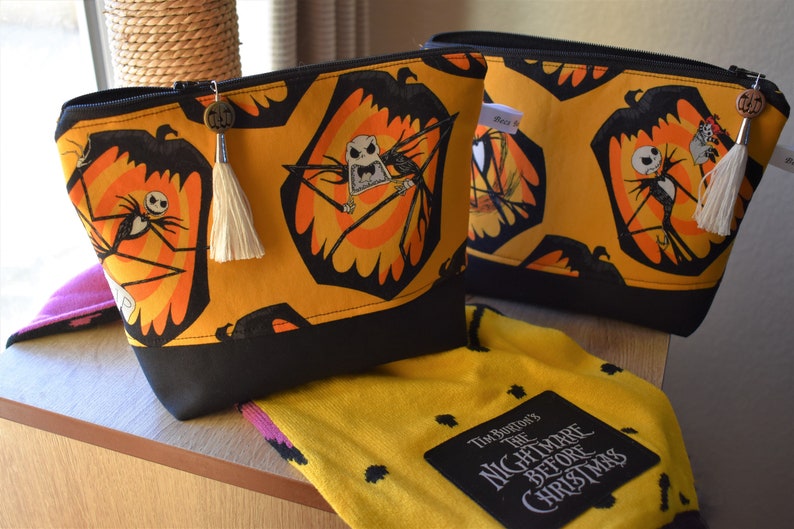 May include: Two yellow and black zippered pouches with a black and white Jack Skellington print. The pouches have a black tassel and a metal charm. A yellow and black fabric with the text "Tim Burton's The Nightmare Before Christmas" is in the foreground.
