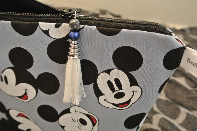 May include: A grey zippered pouch with a Mickey Mouse print. The pouch has a white tassel with a blue and silver bead charm.
