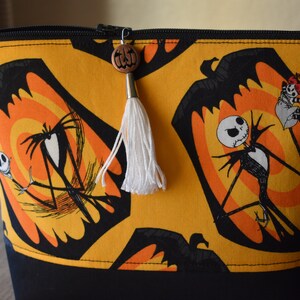 May include: A yellow and black zippered pouch with a repeating pattern of Jack Skellington from The Nightmare Before Christmas. The pouch has a white tassel and a wooden pumpkin charm.