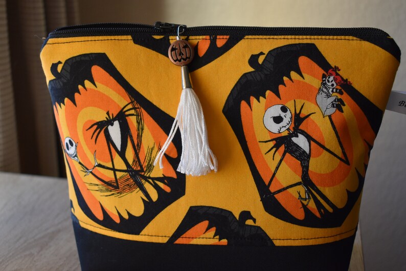 May include: A yellow and black zippered pouch with a repeating pattern of Jack Skellington from The Nightmare Before Christmas. The pouch has a white tassel and a wooden pumpkin charm.