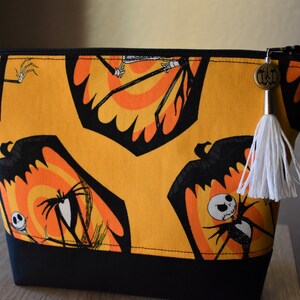May include: A black and orange zippered pouch with a white tassel. The pouch features a repeating pattern of Jack Skellington from The Nightmare Before Christmas.