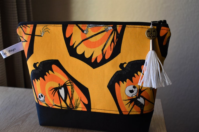 May include: A black and orange zippered pouch with a white tassel. The pouch features a repeating pattern of Jack Skellington from The Nightmare Before Christmas.
