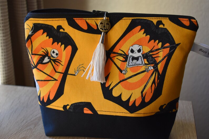May include: A yellow zippered pouch with a black and white print of Jack Skellington from The Nightmare Before Christmas. The pouch has a black zipper and a white tassel with a pumpkin charm.
