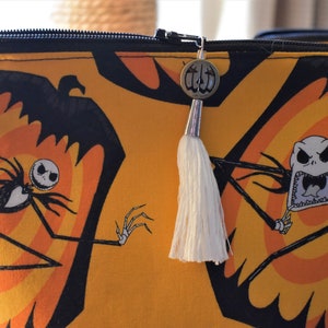 May include: A yellow zippered pouch with a black and orange print of Jack Skellington from The Nightmare Before Christmas. The pouch has a white tassel attached to a silver charm.