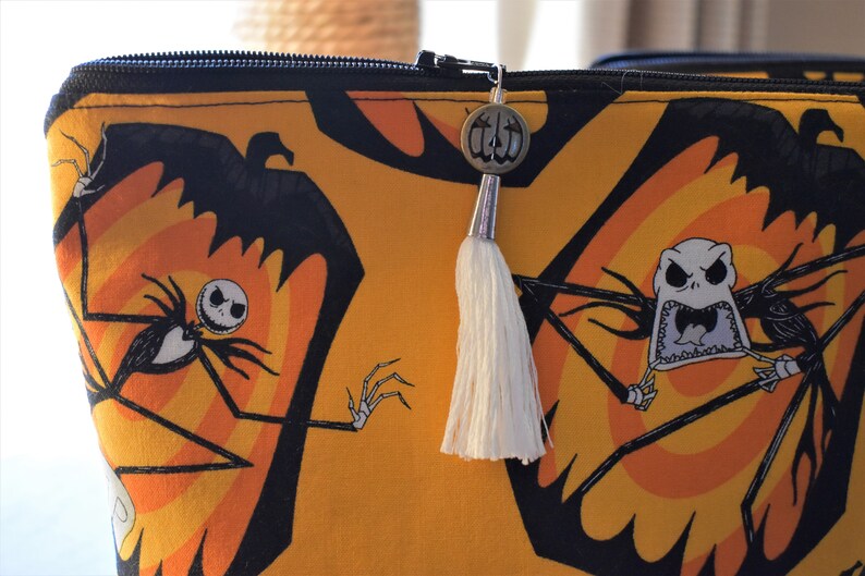 May include: A yellow zippered pouch with a black and orange print of Jack Skellington from The Nightmare Before Christmas. The pouch has a white tassel attached to a silver charm.