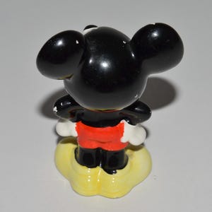 May include: A vintage Mickey Mouse figurine, viewed from the back. The figure has a glossy black head and ears, red shorts, and white gloves. It stands on a yellow cloud-shaped base. A classic Disney collectible.