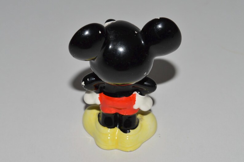 May include: A vintage Mickey Mouse figurine, viewed from the back. The figure has a glossy black head and ears, red shorts, and white gloves. It stands on a yellow cloud-shaped base. A classic Disney collectible.