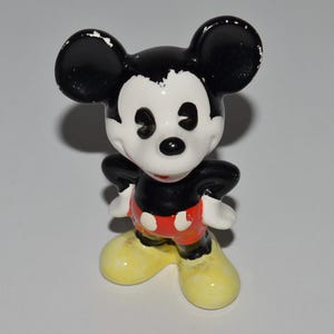 May include: A small, vintage Mickey Mouse figurine. The figure has a black head with large ears, a white face, and a black nose. It wears a black top, red shorts with white buttons, yellow shoes, and white gloves. The figurine is standing with its hands on its hips.