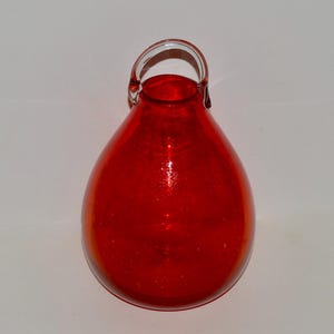 May include: A red glass vase with a clear glass handle. The vase is shaped like a teardrop and has a textured surface.
