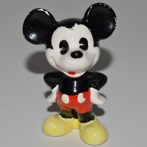 May include: A ceramic Mickey Mouse figurine. The figure has a black head and ears, a white face, black torso, red shorts, white gloves, and yellow shoes. The figure is standing with its hands on its hips.