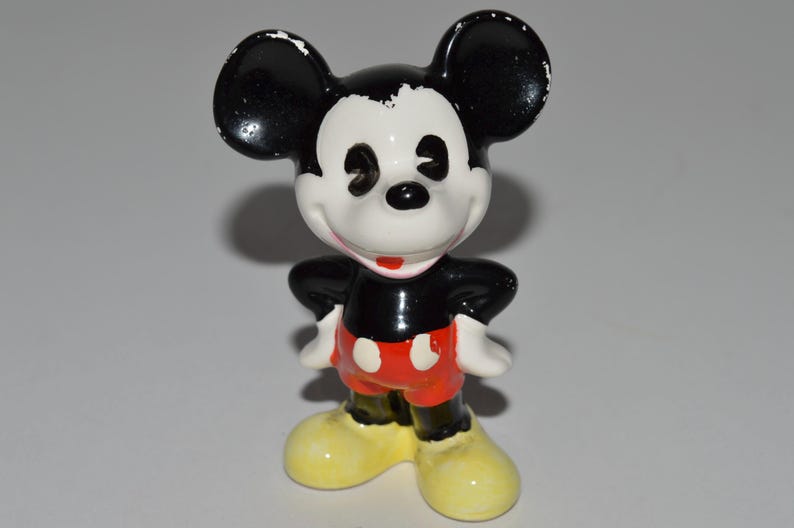 May include: A ceramic Mickey Mouse figurine. The figure has a black head and ears, a white face, black torso, red shorts, white gloves, and yellow shoes. The figure is standing with its hands on its hips.
