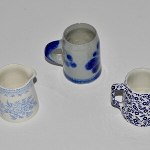 May include: Three ceramic pitchers with blue and white designs. One pitcher has a floral pattern, another has a speckled gray and blue design, and the third has a blue floral pattern. All have handles and spouts.