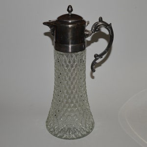 May include: A silver-plated glass pitcher with a clear, textured body and a decorative handle. The pitcher has a silver-plated lid with a small knob on top.