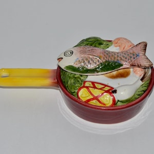 May include: A red ceramic casserole dish with a yellow handle. The dish is decorated with a painted scene of a fish, vegetables, and other food items.