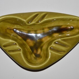 May include: A green ceramic ashtray with a triangular shape and three indented areas for cigarettes. The ashtray has a glossy finish and a brown glaze in the center.