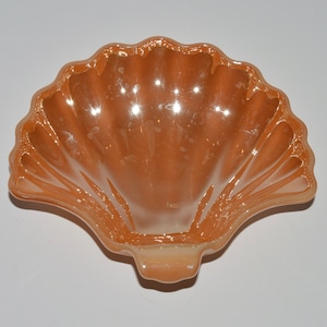 May include: A peach-colored glass shell-shaped dish. The dish has a scalloped edge and a smooth, shiny surface.