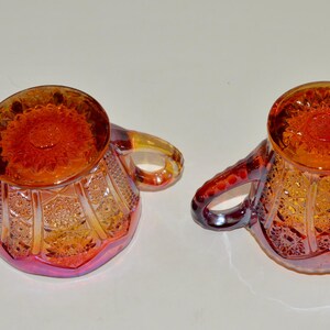 May include: A set of two vintage glass sugar bowls with a cranberry and amber iridescent finish. The bowls have a scalloped edge and a floral design.