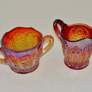 May include: A set of two vintage glass sugar and creamer bowls. The bowls are made of iridescent glass with a red, orange, and yellow color scheme. The bowls have a decorative pattern and are in good condition.