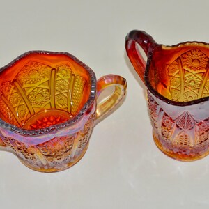 May include: A set of two vintage glass sugar and creamer in a cranberry color with a clear iridescent finish. The set features a detailed geometric pattern.