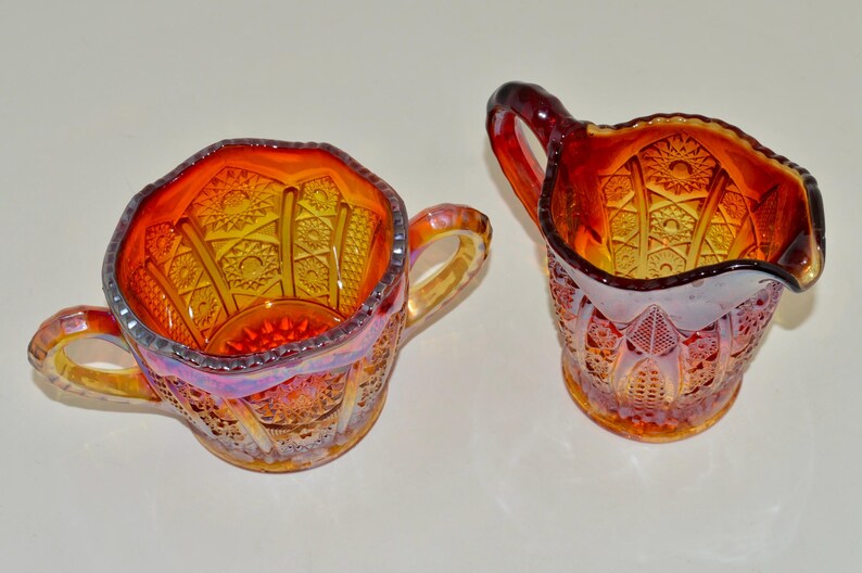 May include: A set of two vintage glass sugar and creamer in a cranberry color with a clear iridescent finish. The set features a detailed geometric pattern.