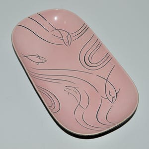 May include: A pink, oval-shaped ceramic dish with black line drawings of fish and wavy lines. The dish has a glossy finish and a slightly raised edge. The design is minimalist and modern, suitable for serving food or as a decorative piece.
