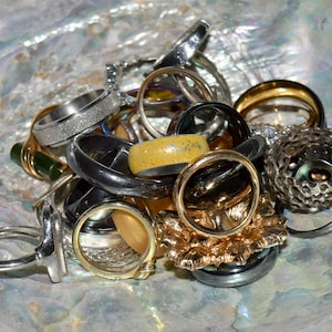 May include: A collection of various rings in gold, silver, and black tones, displayed in a pearlescent shell. The rings feature diverse designs, including textured bands, gemstone settings, and intricate floral patterns, creating a mix of styles.