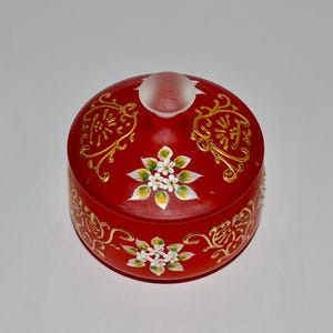 May include: A red glass jar with a gold and white floral design. The lid is frosted glass.