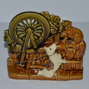 May include: A ceramic planter shaped like a scene with a dog, a cat, and a spinning wheel. The planter is brown and green with white accents. The dog is white with brown spots and is looking at the cat. The cat is brown and is looking at the spinning wheel. The spinning wheel is green and has a brown wooden frame.