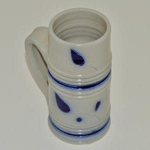 May include: A white ceramic beer stein with a blue and white floral design. The stein has a handle and a wide mouth.