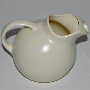 May include: Cream-colored ceramic pitcher with a rounded body and a curved handle. The pitcher has a spout for pouring liquids. The design is simple and elegant, suitable for serving beverages.