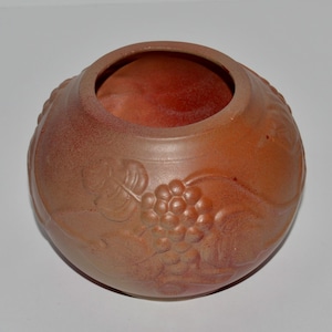 May include: A round, terracotta-colored ceramic vase with a wide opening. The exterior features a raised relief design of grape clusters and leaves. The interior has a reddish hue, and the vase sits on a white surface.