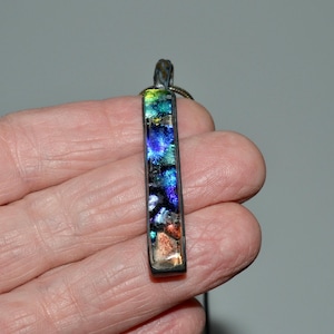 May include: A rectangular pendant with a colorful, abstract design. The pendant features a mix of blues, greens, and orange hues, with a dark border and a silver-toned bail. The pendant is suspended from a black cord.